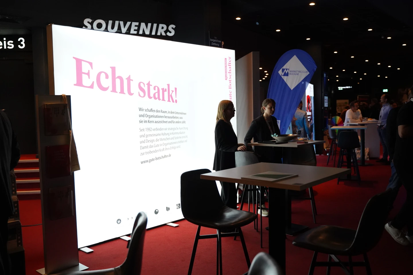 Leuchtende Werbetafel mit der &Uuml;berschrift &bdquo;Echt stark!&ldquo; in pinker Schrift, darunter Textbl&ouml;cke. Zwei Personen an einem Stehtisch, rechts eine weitere Person an einem Stand mit blauer Werbeflagge. Rot beleuchteter Boden, schwarze St&uuml;hle, im Hintergrund der Schriftzug &bdquo;SOUVENIRS&ldquo; und weitere Messe-St&auml;nde.