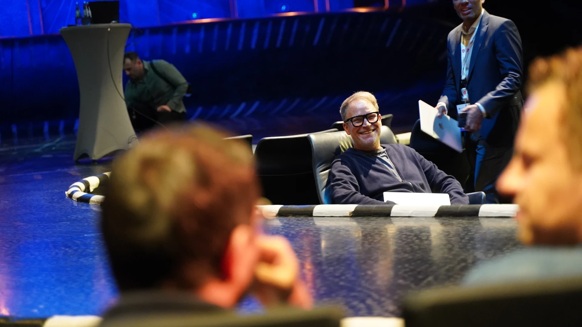 Mann mit Brille und dunkelblauem Pullover sitzt auf einem schwarzen Sessel, l&auml;chelnd. Im Hintergrund ein l&auml;chelnder Mann im Anzug mit Papier in der Hand. B&uuml;hne mit blauen Lichtern und Stehtisch links sichtbar. Unscharfe Personen im Vordergrund.