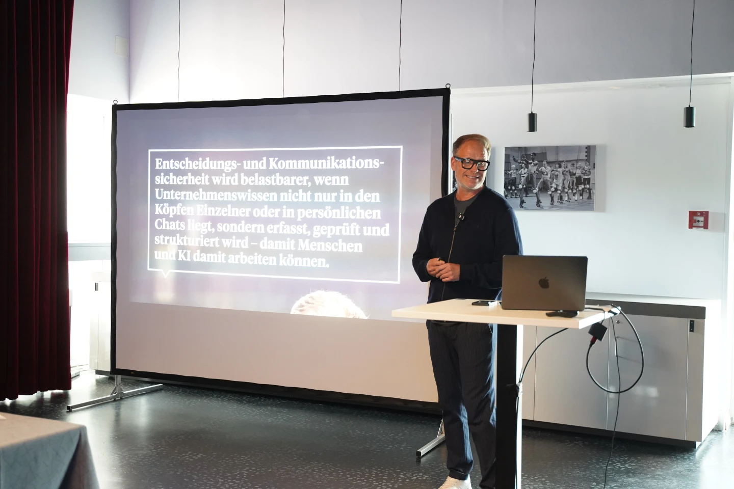 Mann mit schwarzer Brille und dunkler Kleidung steht neben einem wei&szlig;en Stehpult mit schwarzem Laptop. Im Hintergrund eine wei&szlig;e Leinwand mit projiziertem Text: "Entscheidungs- und Kommunikationssicherheit wird belastbarer, wenn Unternehmenswissen nicht nur in den K&ouml;pfen Einzelner oder in pers&ouml;nlichen Chats liegt, sondern erfasst, gepr&uuml;ft und strukturiert wird &ndash; damit Menschen und KI damit arbeiten k&ouml;nnen." Raum mit wei&szlig;en W&auml;nden und roten Vorh&auml;ngen links.