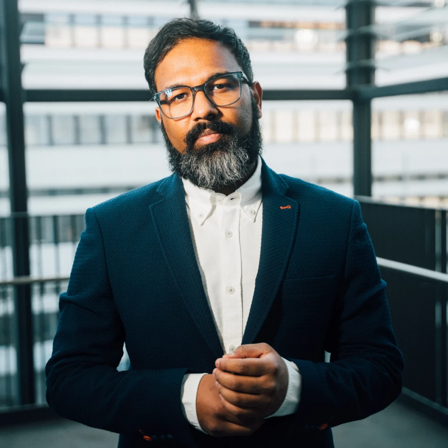 Mann mit Brille, Vollbart und dunklem Anzug mit wei&szlig;em Hemd, steht mit gefalteten H&auml;nden auf einer Terrasse. Im Hintergrund sind Gel&auml;nder und ein modernes mehrst&ouml;ckiges Geb&auml;ude mit gro&szlig;en Fenstern sichtbar. Hell-dunkle Lichtakzente durch Sonneneinstrahlung.