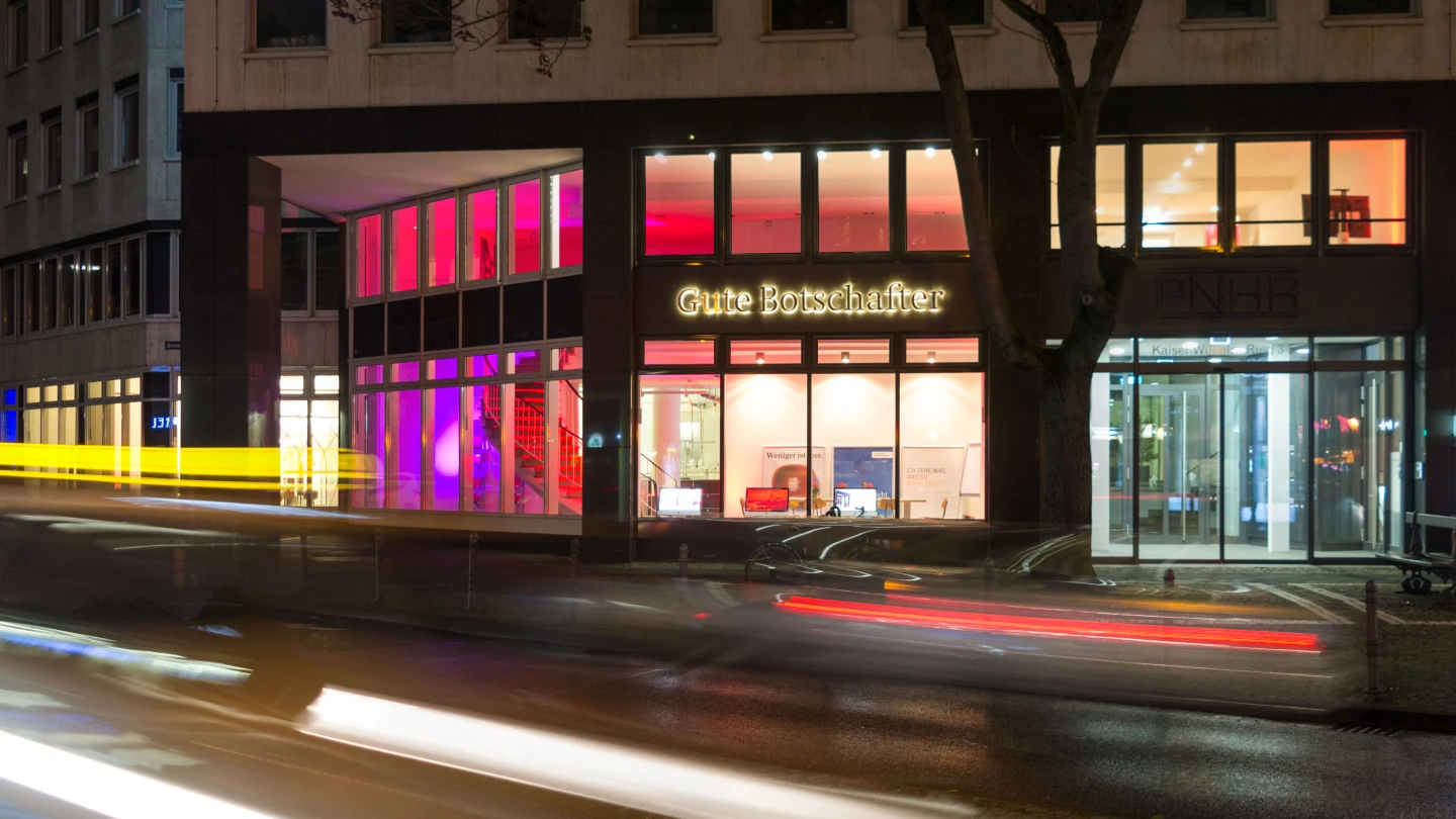 Beleuchtetes Geb&auml;ude bei Nacht mit Schriftzug "Gute Botschafter" im mittleren Bereich der Fassade. Linke Geb&auml;udeseite mit roten und pinken Lichtakzenten, rechts Glaseingang mit Schriftzug "ANHB". Vor dem Geb&auml;ude Lichtspuren von vorbeifahrenden Autos.