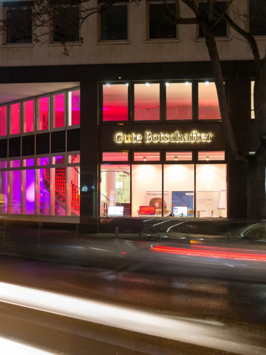 Beleuchtetes Geb&auml;ude bei Nacht mit Schriftzug "Gute Botschafter" im mittleren Bereich der Fassade. Linke Geb&auml;udeseite mit roten und pinken Lichtakzenten, rechts Glaseingang mit Schriftzug "ANHB". Vor dem Geb&auml;ude Lichtspuren von vorbeifahrenden Autos.