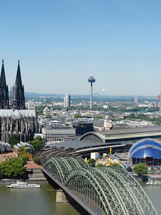 Luftaufnahme einer Stadt mit dem K&ouml;lner Dom, einer gotischen Kathedrale mit zwei markanten T&uuml;rmen, in der Bildmitte. Rechts daneben der Hauptbahnhof und die Lanxess-Arena. Im Vordergrund die Hohenzollernbr&uuml;cke &uuml;ber den Rhein, unter der zwei Ausflugsschiffe fahren. Im Hintergrund der Fernsehturm, moderne Geb&auml;ude und eine weitl&auml;ufige Landschaft.